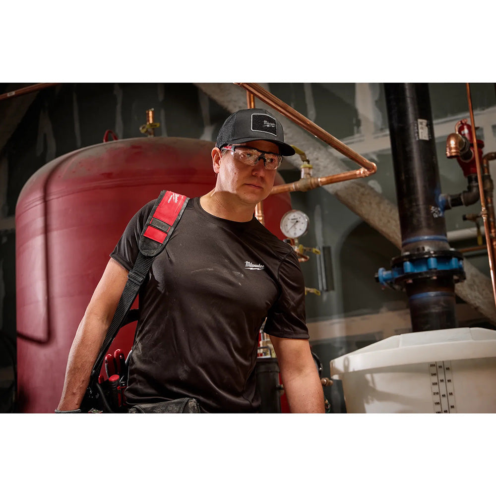 Person wearing a black shirt and cap in an industrial setting with pipes and equipment.